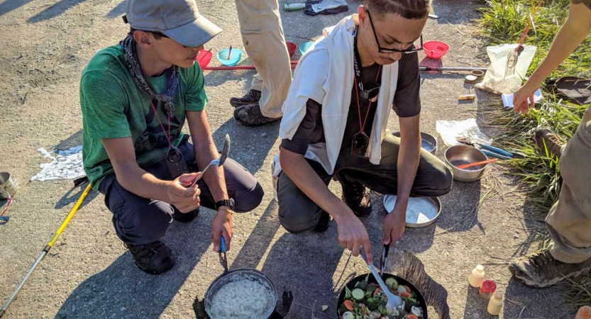 Two students prepare food using camping stoves.